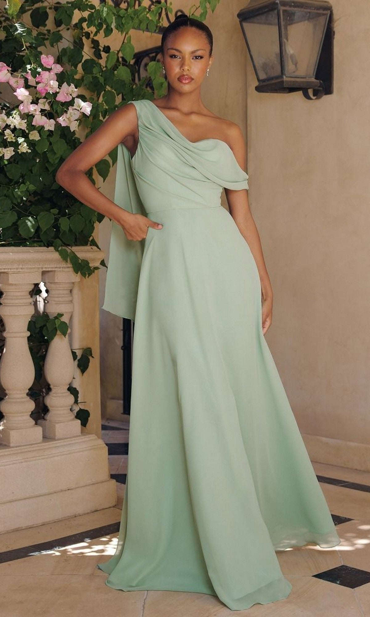 A woman stands outdoors in the Amoris T716 Asymmetric Long A-Line Prom Dress, featuring a light green one-shoulder design with draped fabric—an elegant and affordable choice for proms or any special occasion.